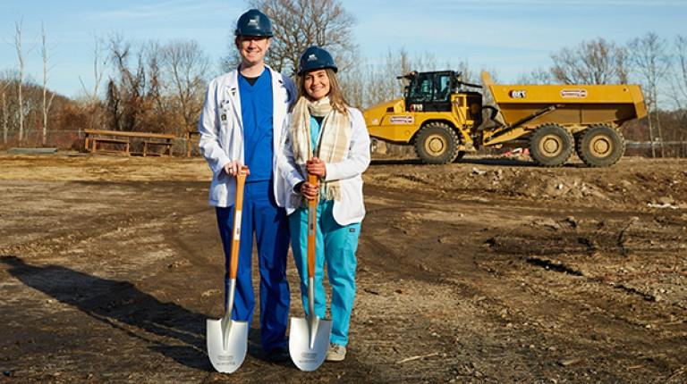 Two UNE COM students pose with shovels, the construction site in the background